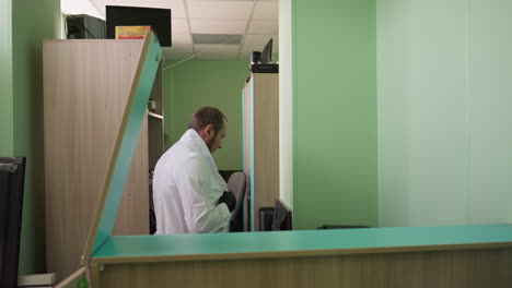 a side view of a man putting on his lab coat in a professional setting, the man is adjusting his collar while standing near a desk in a green-walled laboratory
