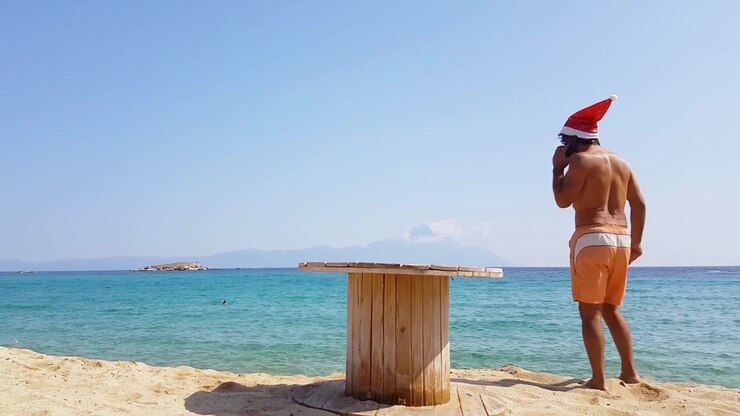 Uomo in costume da bagno e cappello da Babbo Natale che parla al telefono su una spiaggia tropicale. Concetto di Natale e Capodanno.