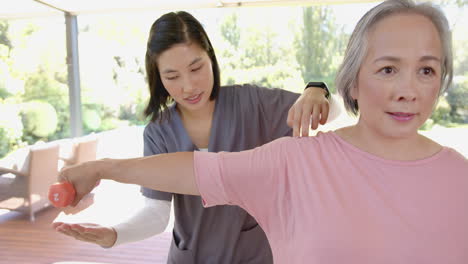 assisting senior asian woman with arm exercise, physical therapist using dumbbell at home