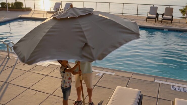 Padre e figlio spingono la manovella, alzando un ombrellone da patio sul bordo piscina per l'ombra, con l'acqua illuminata dal sole.