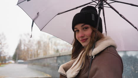 young girl looking at camera smiling, holding umbrella, wearing knit cap, brown shearling jacket, with cheerful expression during light snowfall in a soft, autumn setting