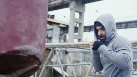 side view of caucasian man in sportswear hitting a punching bag outdoors an abandoned factory on a cloudy morning