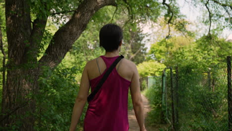 woman wearing pink tanktop walking through a forest trail next to a fence on a sunny summer day
