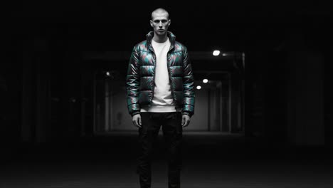a stylish male figure in a black and colorful puffer jacket stands in a dark industrial setting, creating an intriguing contrast with the surroundings