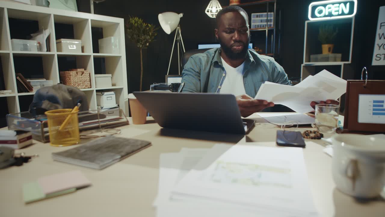 Focused afro man checking data from laptop screen with documents indoors.