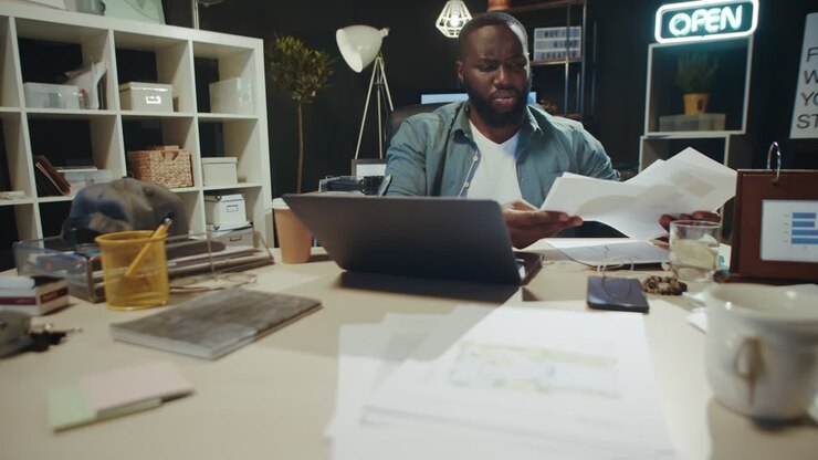 Focused afro man checking data from laptop screen with documents indoors.