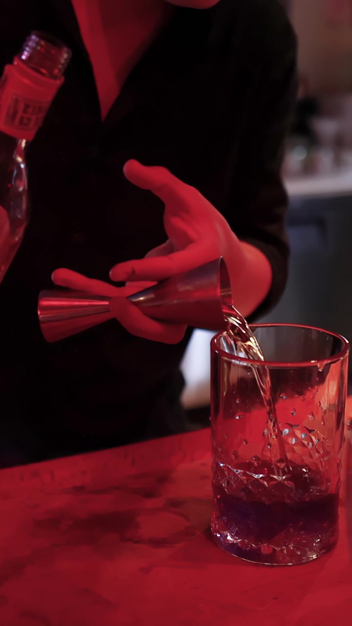 Un barista versa liquore con un jigger in un mixing glass sotto un'illuminazione rossa suggestiva, vista ravvicinata