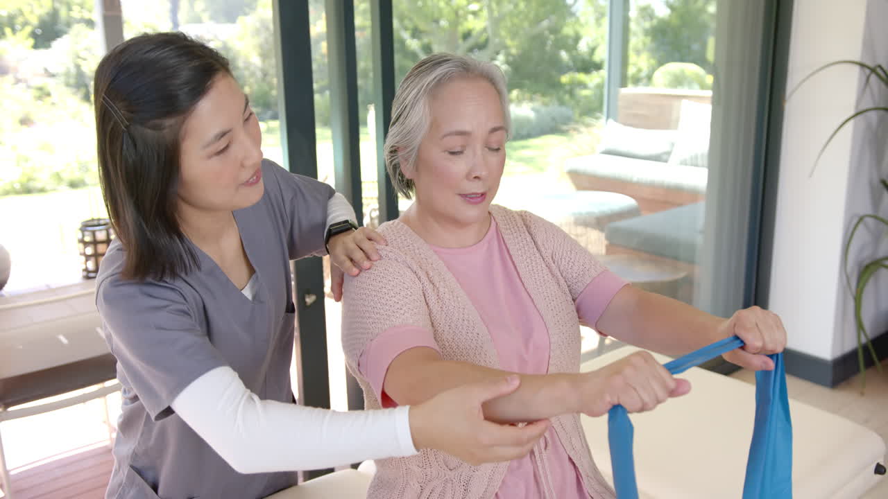 Assisting senior woman with resistance band, physical therapist guiding exercise at home