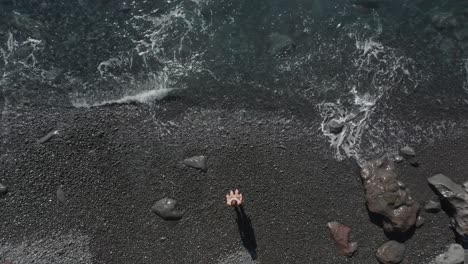 woman in bikini walking and stopping at water edge of waves splashing on pebble beach, top down