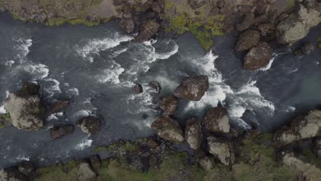 Toma-Aérea-De-Arriba-Hacia-Abajo-De-Un-Hermoso-Río-Y-Una-Cascada-En-Islandia-En-Un-Día-Oscuro-Y-Brumoso-Frente-A-Los-Acantilados-Y-Rocas-Verdes-Y-Cubiertos-De-Musgo