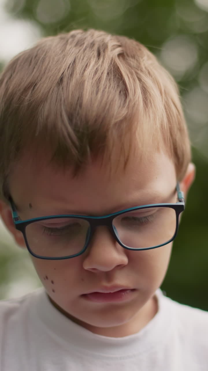un niño con gafas parece abatido, mirando hacia abajo con una expresión triste, el fondo está borroso, mostrando vegetación