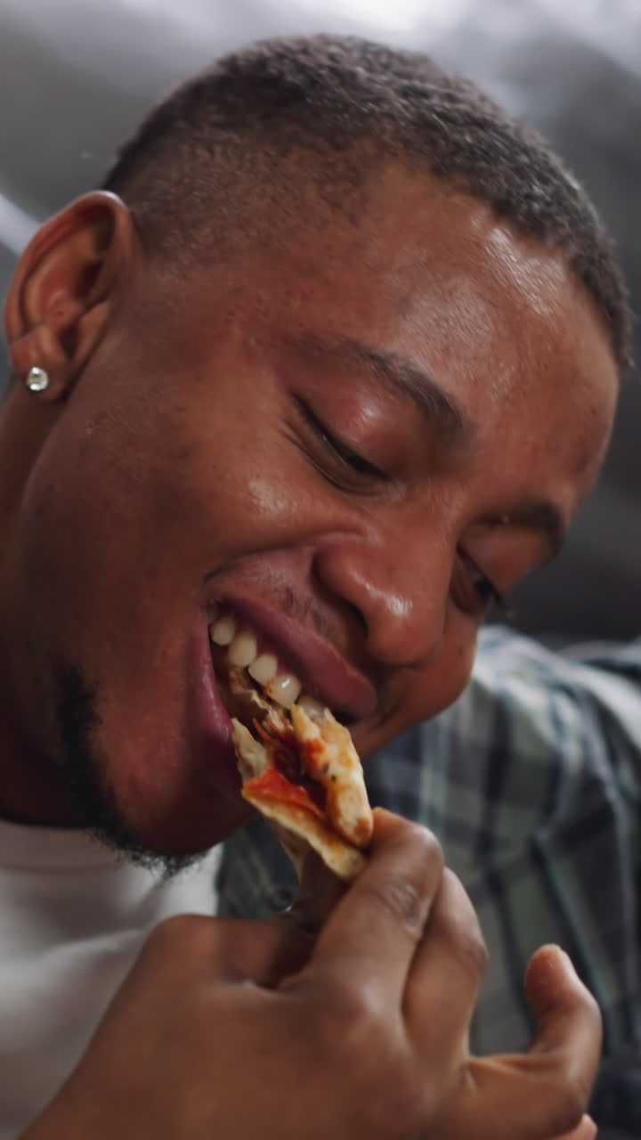 Smiling guy with earring bites pizza slice leaning on sofa covered with foil sheet closeup. Black man in shirt has lunch during home repairing