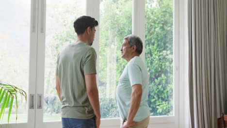 senior man discussing with son while standing by window in living room at home