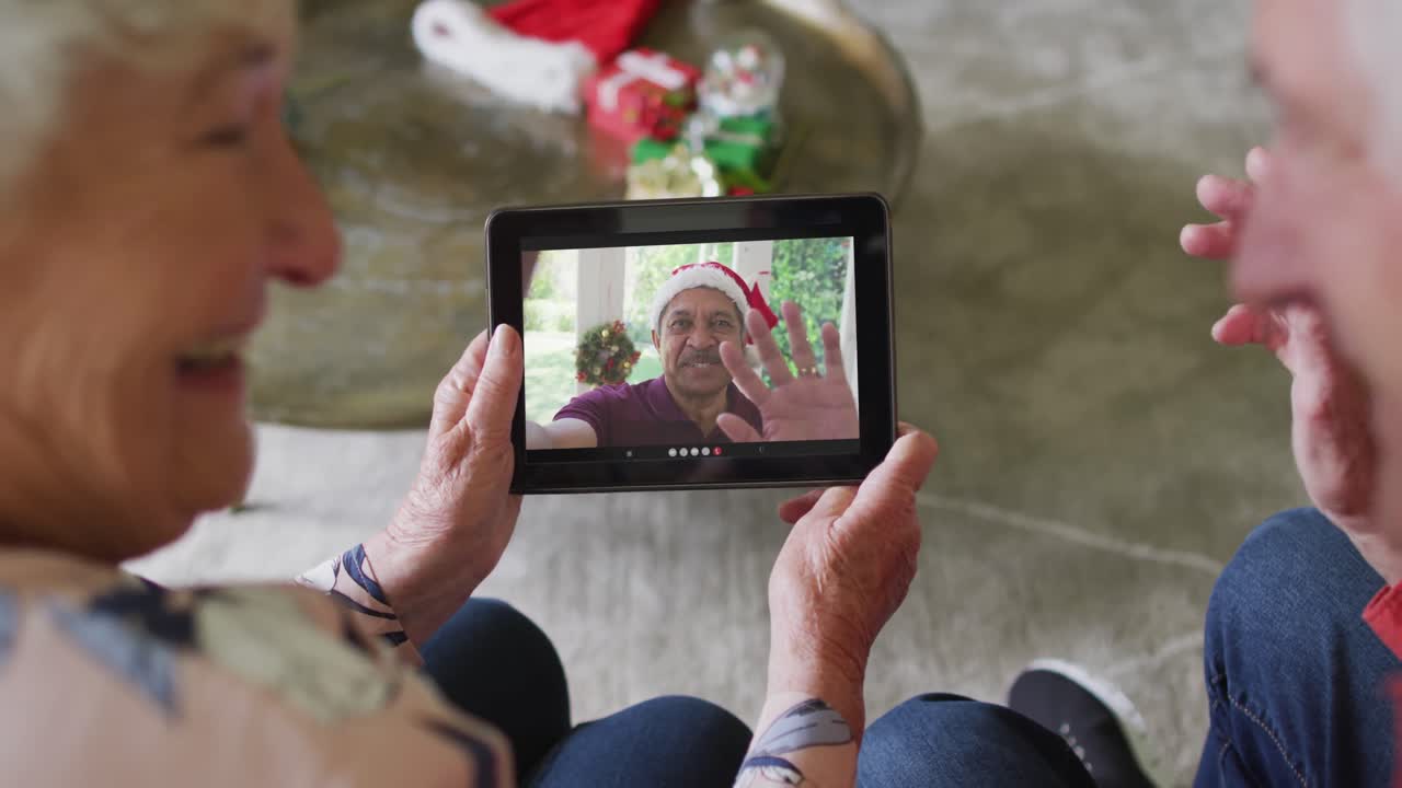 una pareja caucásica mayor sonriente usando una tableta para una videollamada de navidad con un hombre en la pantalla
