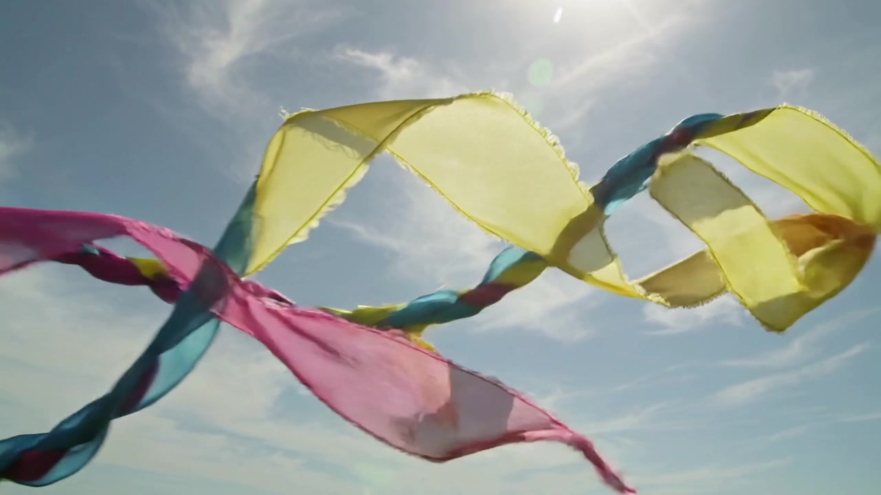 Fitas translúcidas rosa, azul e amarelas torcendo-se, formando um núcleo trançado contra céu claro, impulsionadas pelo vento.