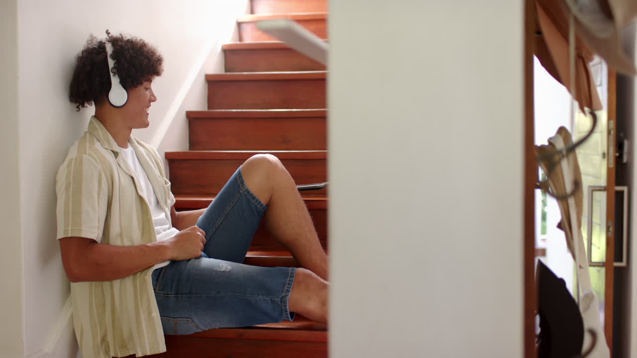 Listening to music with headphones, young man relaxing on staircase steps, at home