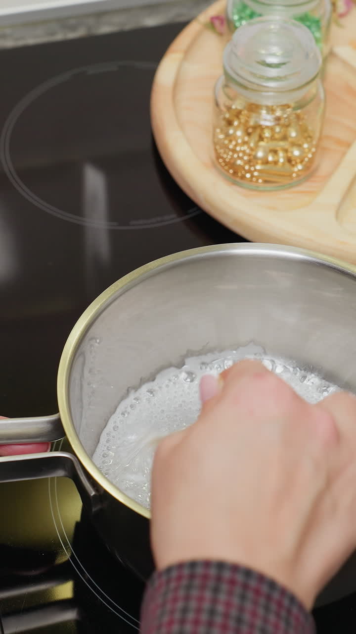 주방 위에 있는 비에 설탕을 는 사람의 근접한 손으로 보는 모습, 다른 색의 구슬이 근처의 나무 트레이에 어져있는 동안, 주방 장면은 설탕 시럽을 조리하는 데 초점을 맞추고 있습니다.