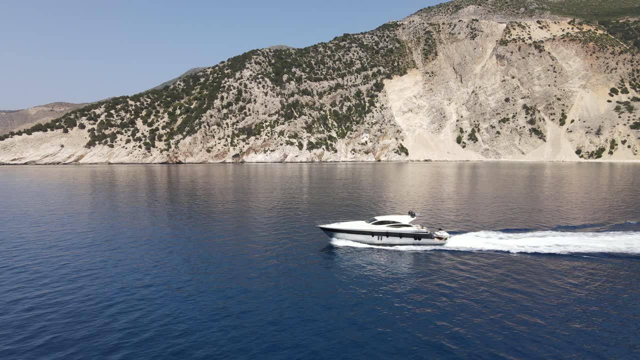 Barca bianca lussuosa naviga lungo la costa greca, inquadratura aerea che si allontana.
