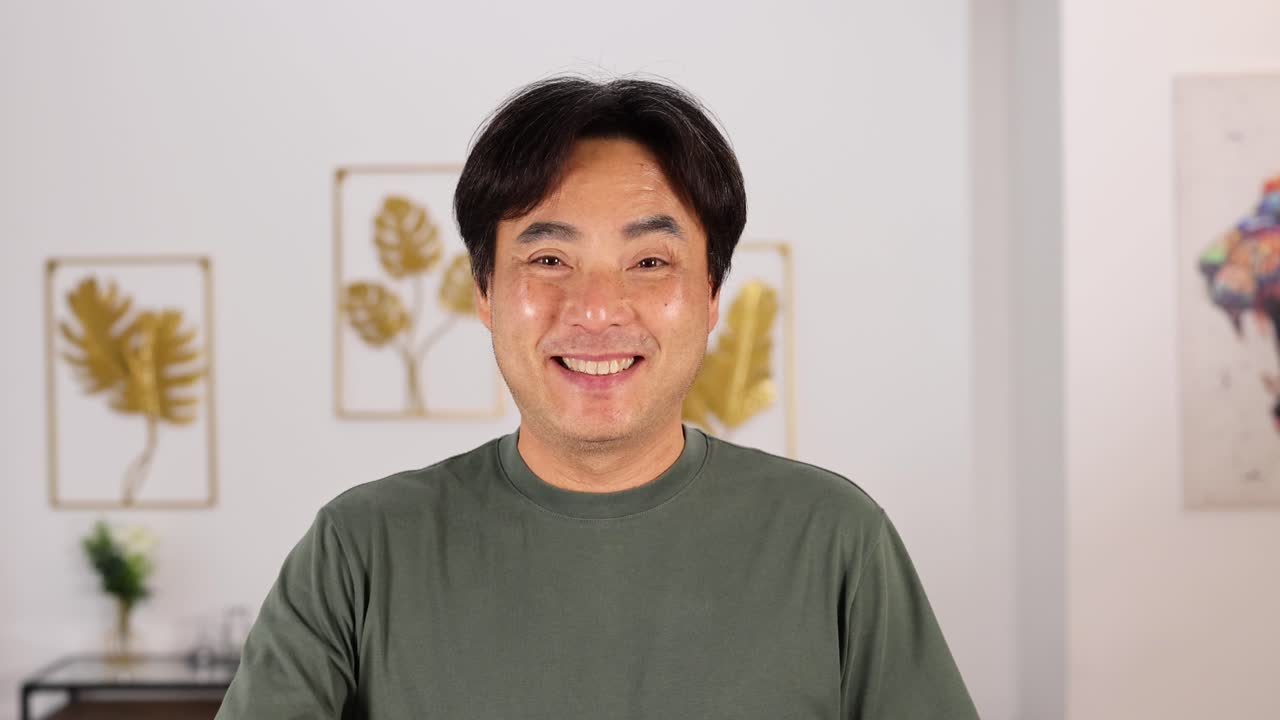 Middle-aged Asian man smiles and makes an OK hand gesture in a well-lit, modern living room with framed art and soft natural lighting