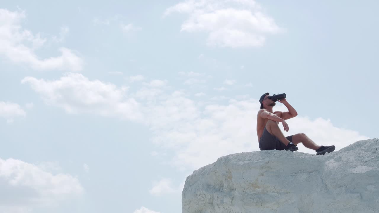 A male athlete sitting on a mountain drinks water from a bottle
