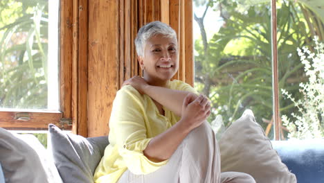 Portrait-of-happy-senior-biracial-woman-sitting-on-couch-at-window-at-home,-slow-motion