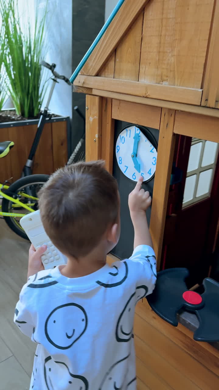Little boy stands at the small wooden play house. Happy kid touches the clock on the wall. High angle view. Vertical video.