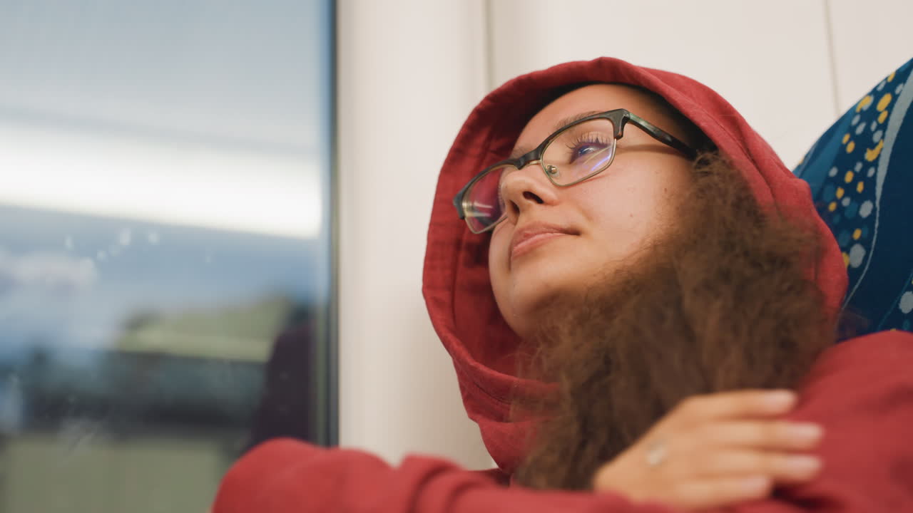 Wearing red hoodie on evening train, pulling hood over head and closing eyes against motion commuter returning home after late shift, soft window reflections, muted city lights, tired expression, hand