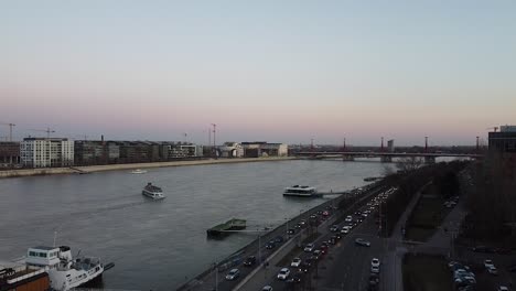 Vista-Aérea-Del-Tráfico-Al-Atardecer-Con-Un-Barco-A-La-Deriva-En-El-Río-Danubio-En-Budapest-Con-La-Ciudad-Al-Fondo