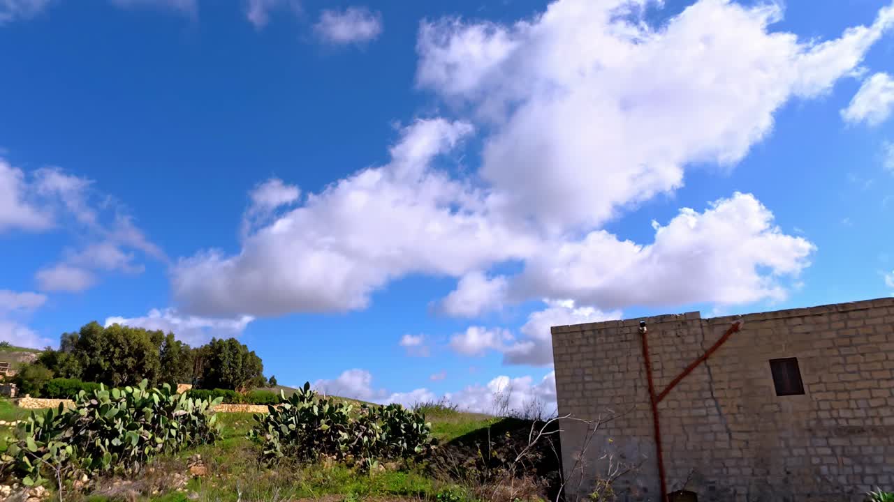 Paisaje primaveral mediterráneo vibrante en Gozo, Malta, con exuberantes colinas verdes en terrazas bajo un cielo azul brillante con nubes cúmulos blancas y esponjosas que muestran un clima soleado y nopal.