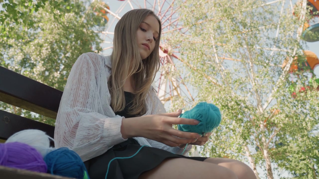 Primer plano de una mujer joven sentada al aire libre en un banco de parque de atracciones, tejiendo activamente con hilo azul. La mujer lleva una blusa de encaje blanca y una falda negra mientras varias bolas de hilo de colores reposan.