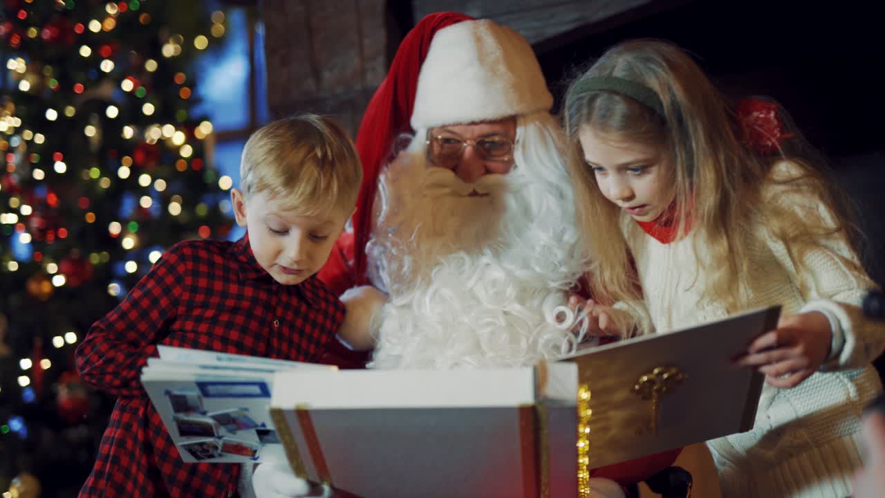 Curious boy and girl looking in album together with Santa Claus. Santa showing photo album to kids on the background of Christmas tree. Close-up.
