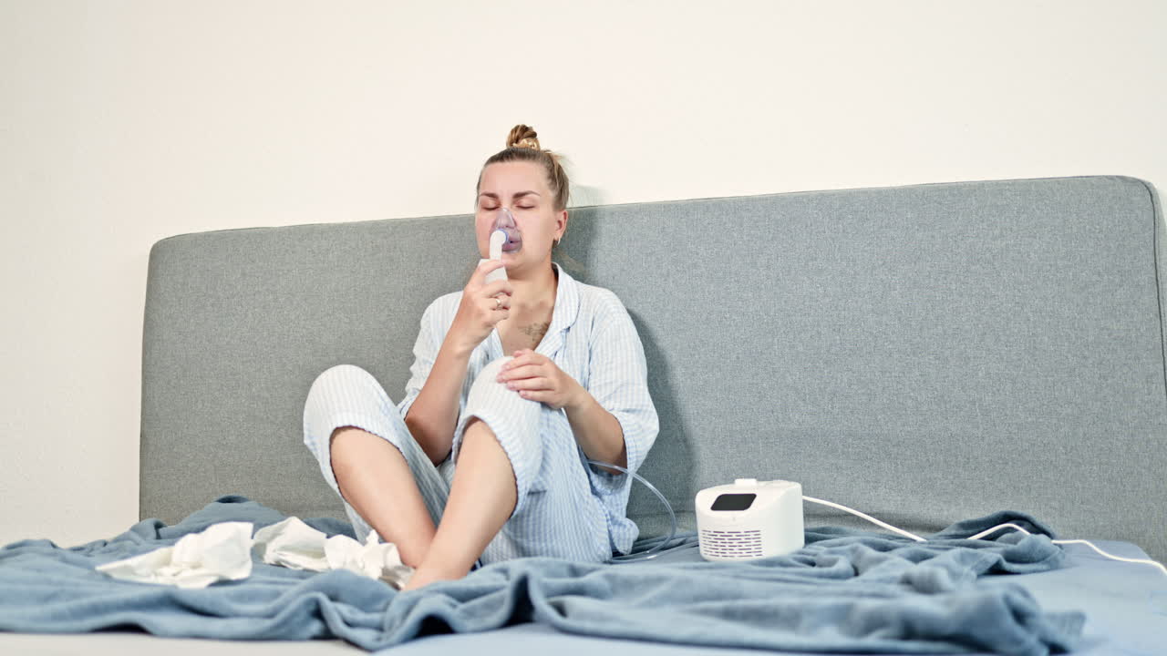 Donna malata che tossisce a casa, seduta sul letto e che respira con un nebulizzatore. Donna che cura difficoltà respiratorie con un inalatore. Automedicazione a casa.