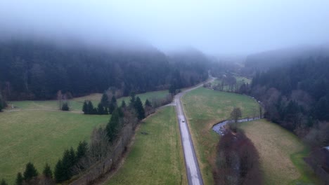 Disparo-De-Un-Dron-Siguiendo-Un-Automóvil-Distante-Sobre-Un-Etéreo-Paisaje-Montañoso-Con-Niebla-Y-Nubes-Bajas