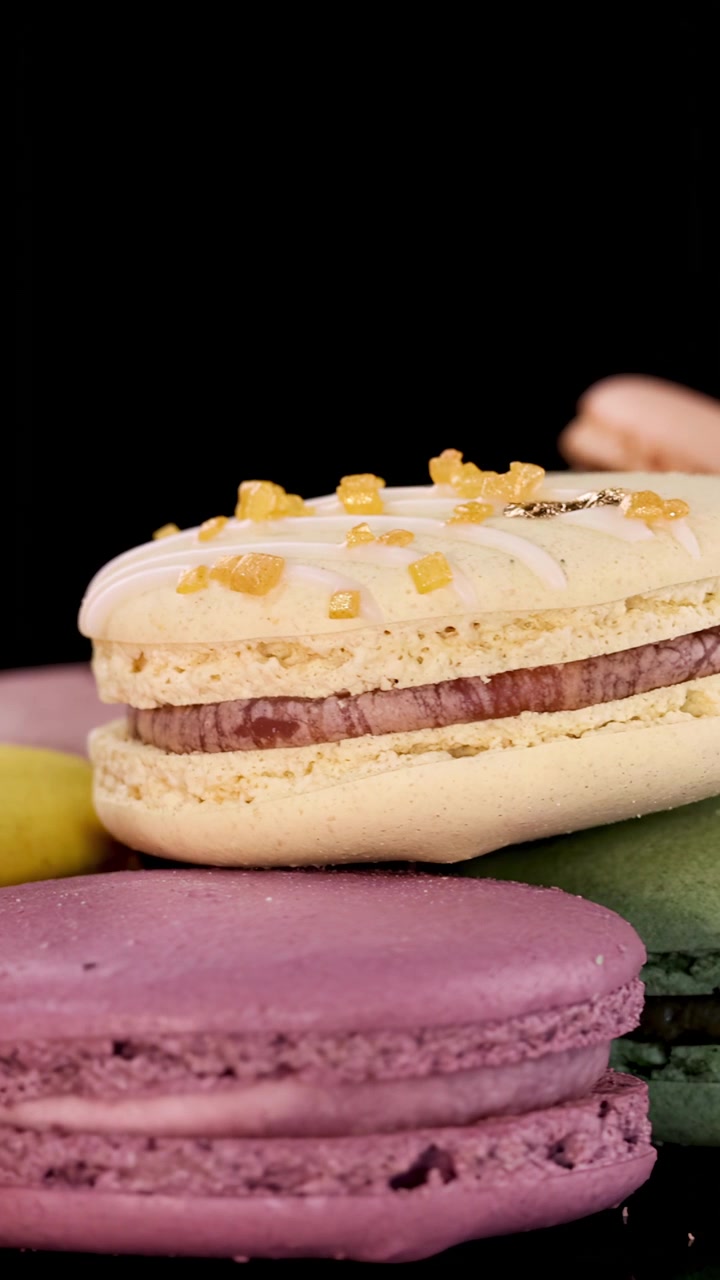 Assorted pastel macarons rotate smoothly on a glossy black surface under bright, even lighting, highlighting texture, color, and detail in a bakery setting