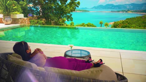 a woman with a sexy evening dress on a trip sitting on a sofa by the pool drinking and looking at the sea