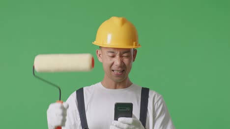Close Up Of Asian Man Painter Wearing Safety Helmet Looking At Smartphone Then Screaming Goal Celebrating While Standing In The Green Screen Background Studio