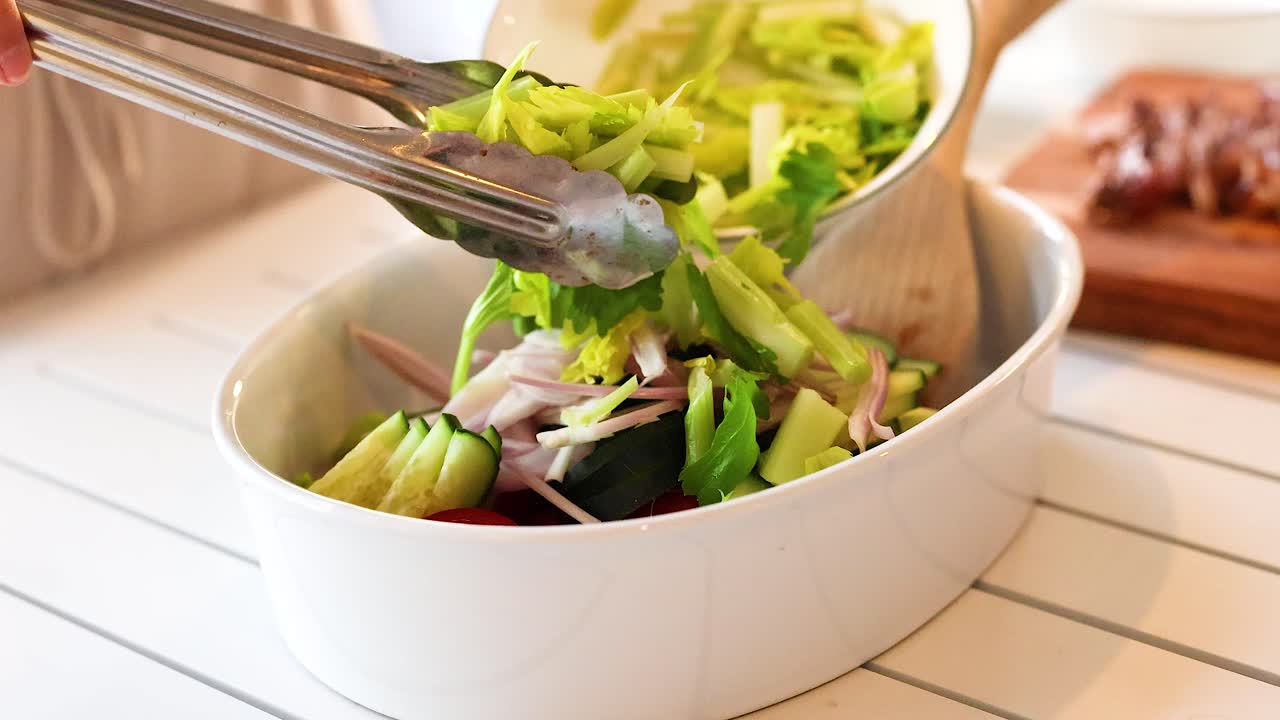 A vibrant salad is assembled with fresh vegetables using tongs in a bright kitchen setting