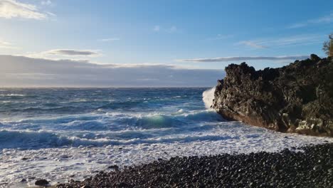amanecer sobre una playa de guijarros negros con olas espumosas golpeando la costa, cielo azul de verano, nubes blancas, cámara lenta