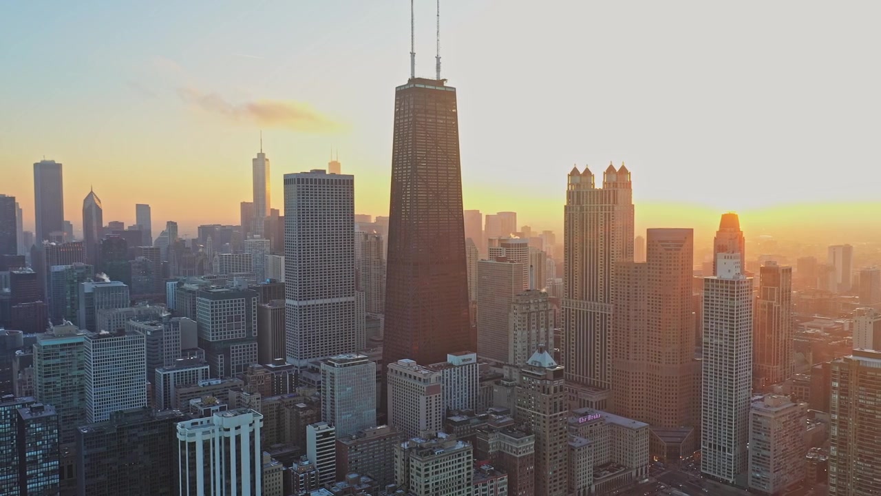Filmische Luftaufnahmen, die Chicagos dichte Skyline und hoch aufragende Stadtarchitektur zeigen, gebadet in sattem goldenem Licht während eines leuchtenden Abendsonnenuntergangs