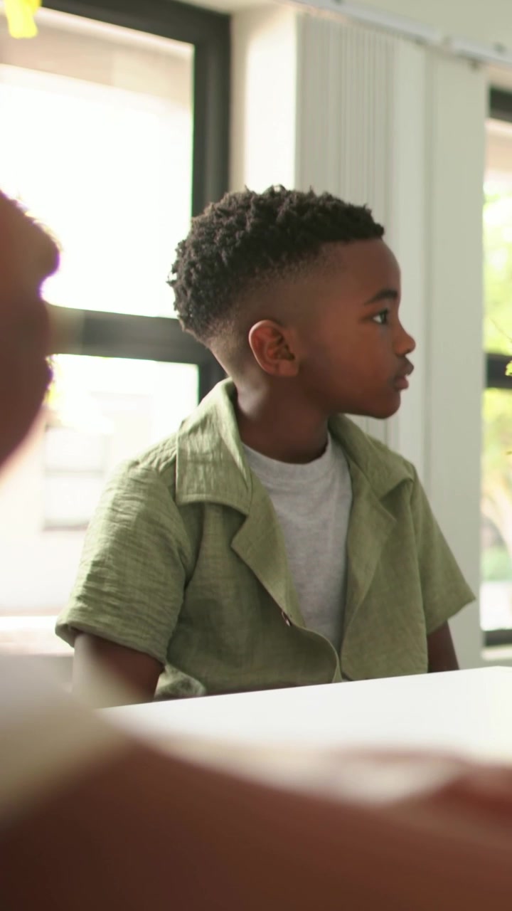 Vertical video: Listening kid in olive tee holding gaze toward offscreen voice at home table, plant