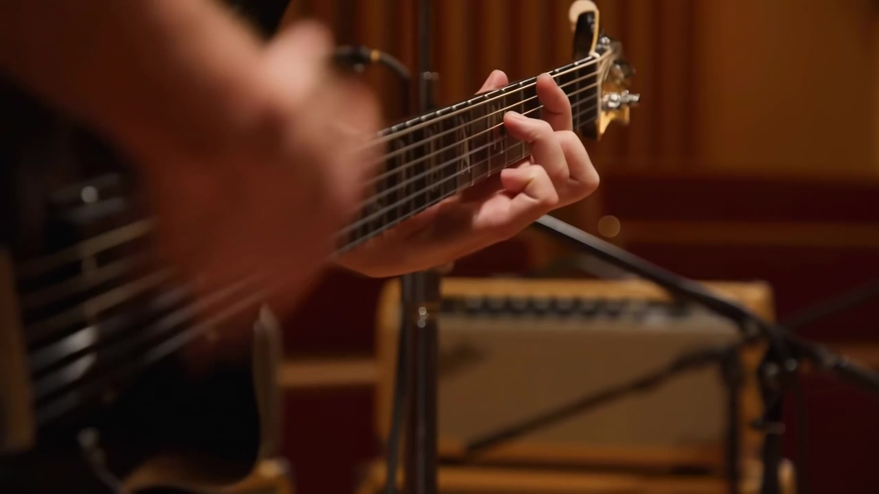 Dettaglio orizzontale ravvicinato delle mani di un chitarrista che suona una chitarra elettrica in uno studio, la mano sinistra forma accordi sulla tastiera vicino alla paletta mentre la mano destra...