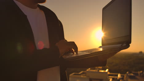 A-male-freelance-programmer-sits-on-a-skyscraper-roof-with-a-laptop-and-beer-typing-code-on-a-keyboard-during-sunset.-Remote-work