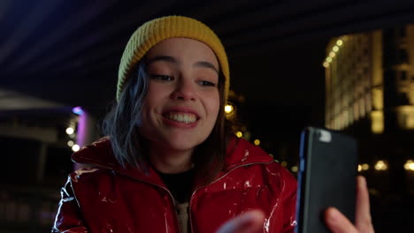 Cheerful-woman-using-cellphone-for-video-call-on-city-street-at-night.