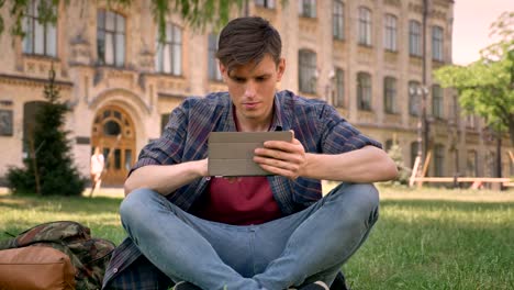 joven está sentado en la hierba en el parque, viendo páginas en internet en la tableta, viendo en la cámara, concepto de relajación, concepto de comunicación, construyendo en el fondo