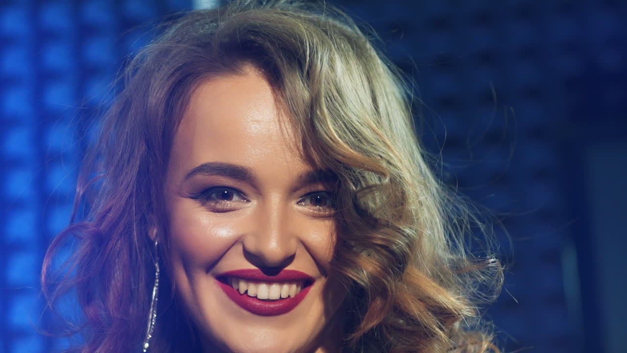 Portrait of a smiling singer with long hair and bare shoulders posing the camera on blue background. Beautiful woman with long earings in studio. Close-up