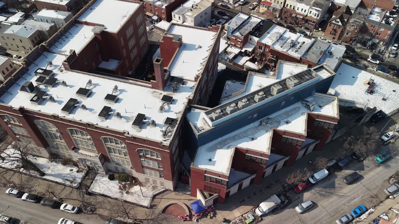 High angle aerial perspective flying backwards away from a brick school building and intersection, revealing a sprawling urban neighborhood with snow covered rooftops on a bright winter day
