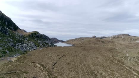 Lowering-aerial-moves-forward-through-moorland-and-rocky-landscape-towards-a-small-remote-loch