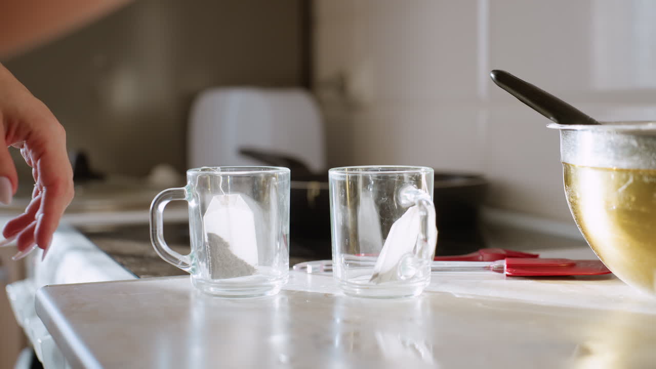 Close up of tea bags being dropped into empty glass cups on kitchen counter with sunlight shining across surface, warm atmosphere of tea preparation