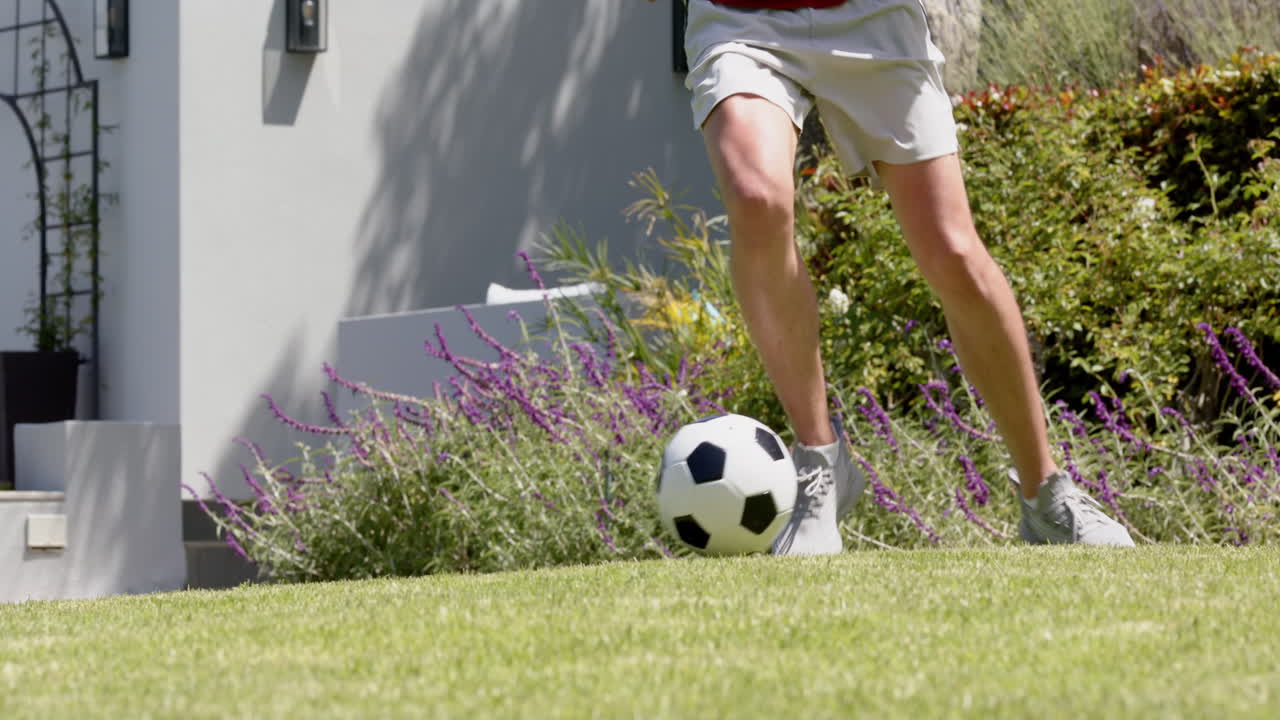 sección baja de hombre biracial practicando habilidades de fútbol en el jardín soleado, cámara lenta