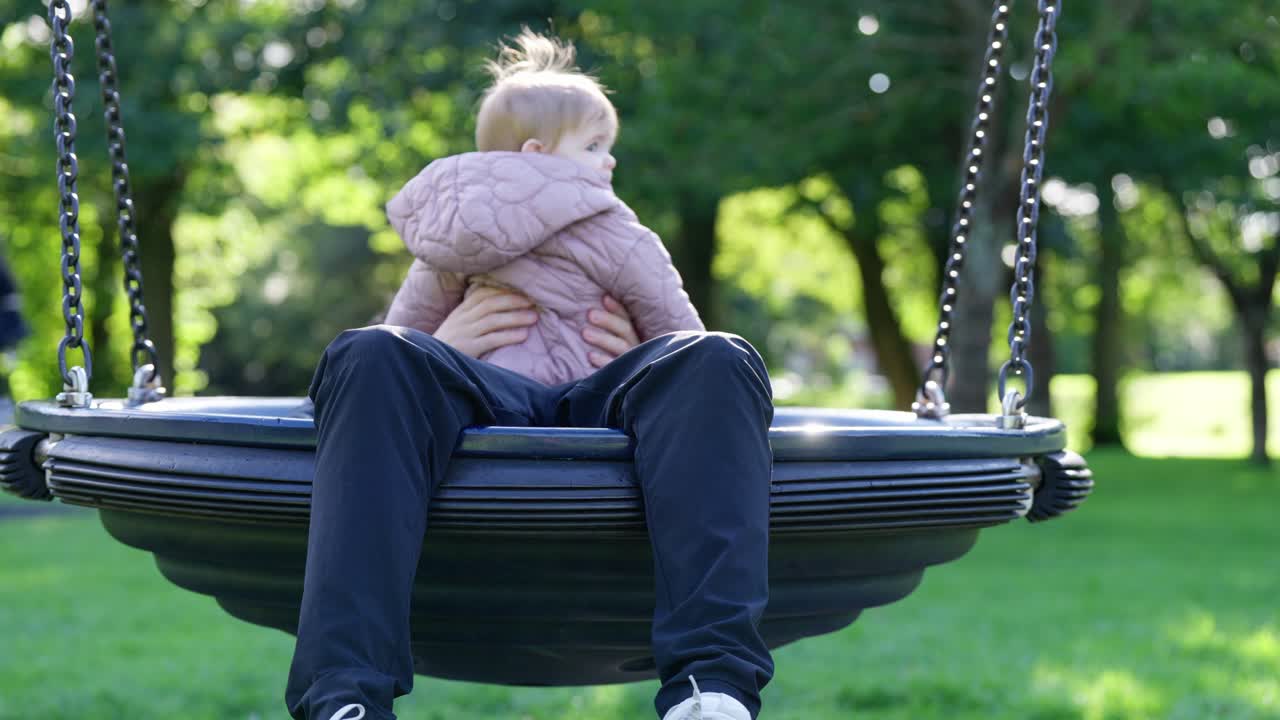 Un giovane padre fa delicatamente dondolare la sua bambina su un'altalena in un parco in Scozia, condividendo un momento tranquillo e gioioso di legame, cura e connessione familiare all'aperto, immerso nel verde della natura.